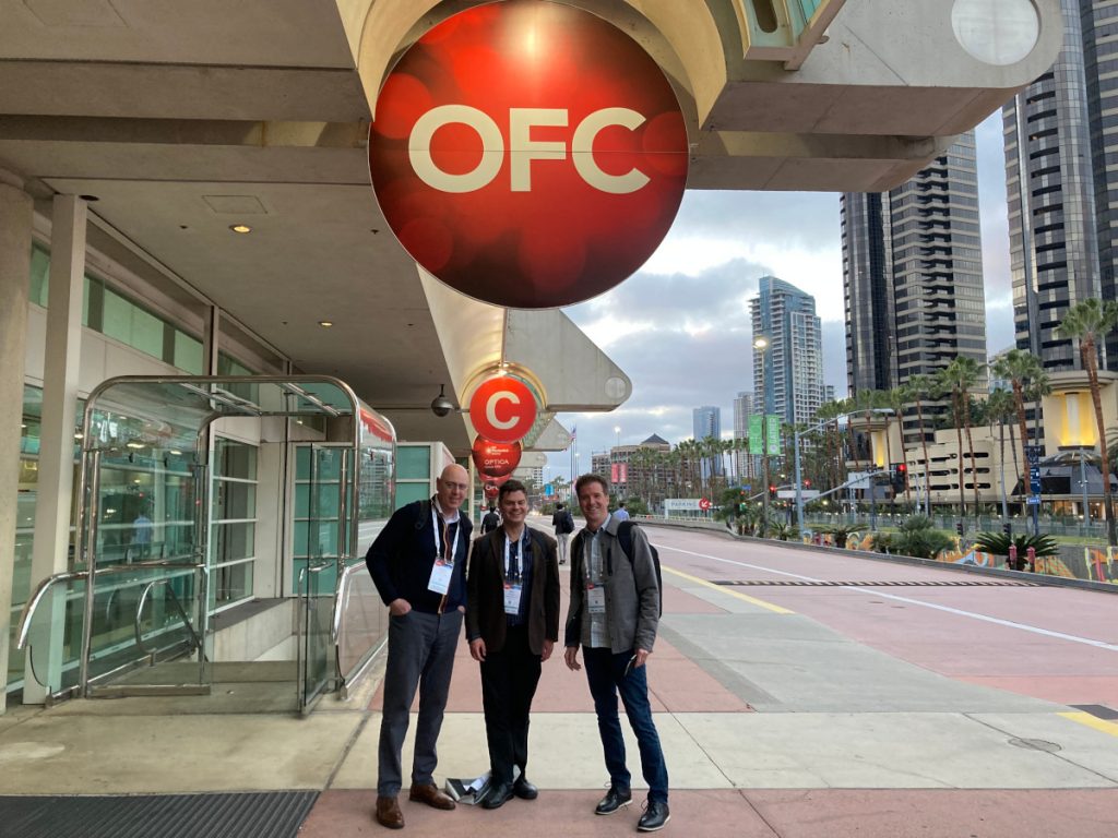 Gerard, Lewis, and David are at OFC in San Diego. They're outside the venue location with the city on the right and the building on the left.