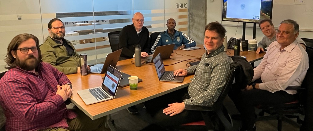 The team gathered around a table with Marion the video call. L-R Kevin, Scott, Gerard, Delwin, Lewis, David, and Vijay