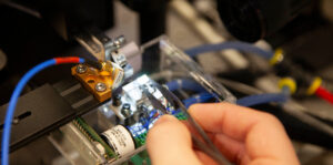 Image shows a close up of a chip being put on a probe station, Scott is holding tweezers which have the chip between them