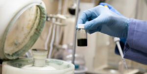 Delwin's hand in a latex glove holds OEO materials in a small glass vial near a machine under the fume hood in NLM's wet lab